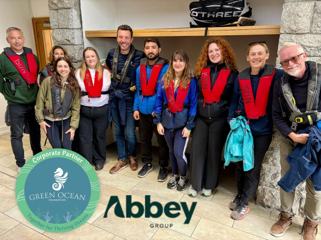 Group of people smiling and posing for a photo with life jackets and outdoor clothes on. Graphic of Green Ocean Corporate Partner logo and Abbey Group logo