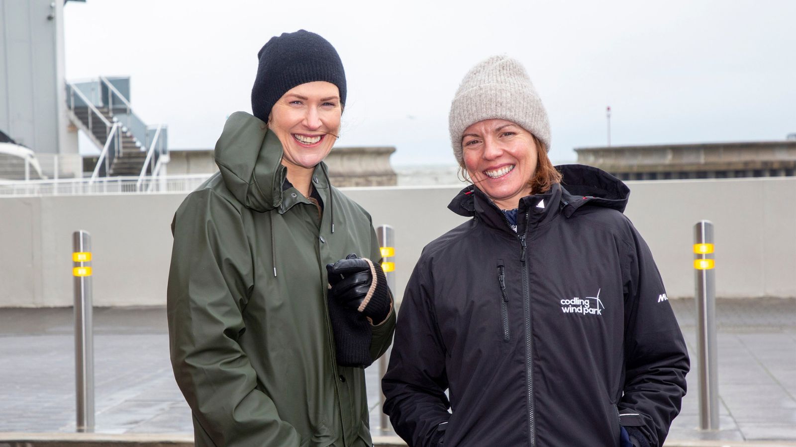 Two women smiling and wearing rain jackets and woolly hats in front of a harbour area