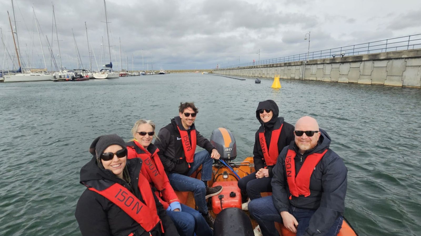 Image of group of people wearing sunglasses, waterproof jackets and life jackets sitting on a speedboat in a marina