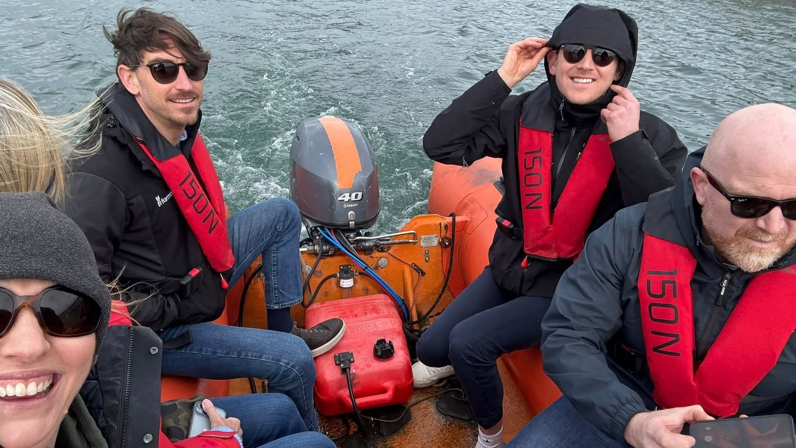 Image of group of people wearing sunglasses, waterproof jackets and life jackets sitting on a speedboat in a marina