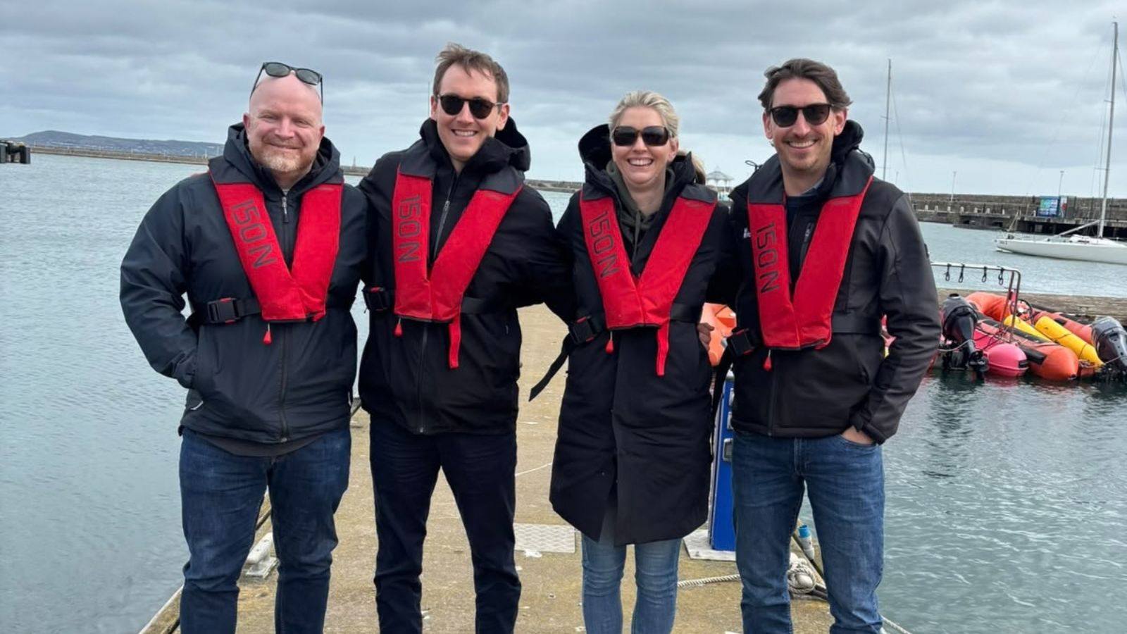 Four people wearing sunglasses, jackets and life jackets, smiling and standing on a pontoon my the sea