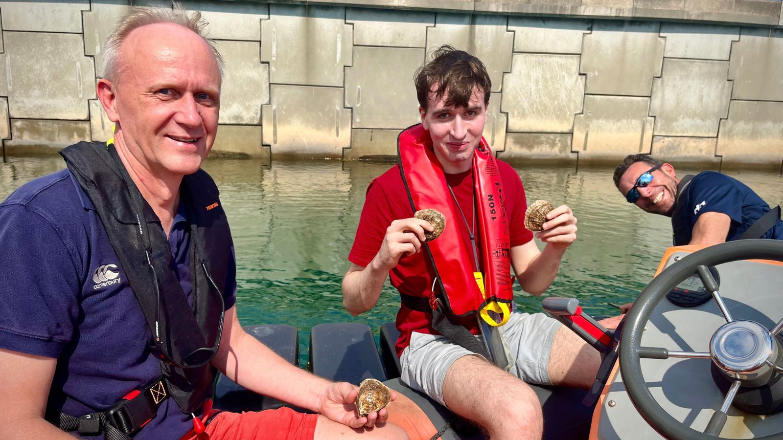 Three men on a rib boat in shorts and smiling holding native oysters