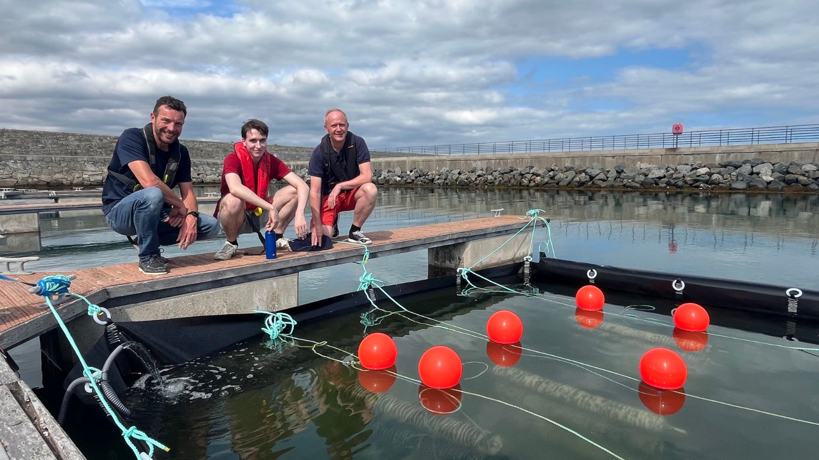 Three men crouching down on a pontoon at sea, beside a temporary spawning pond in a marina.