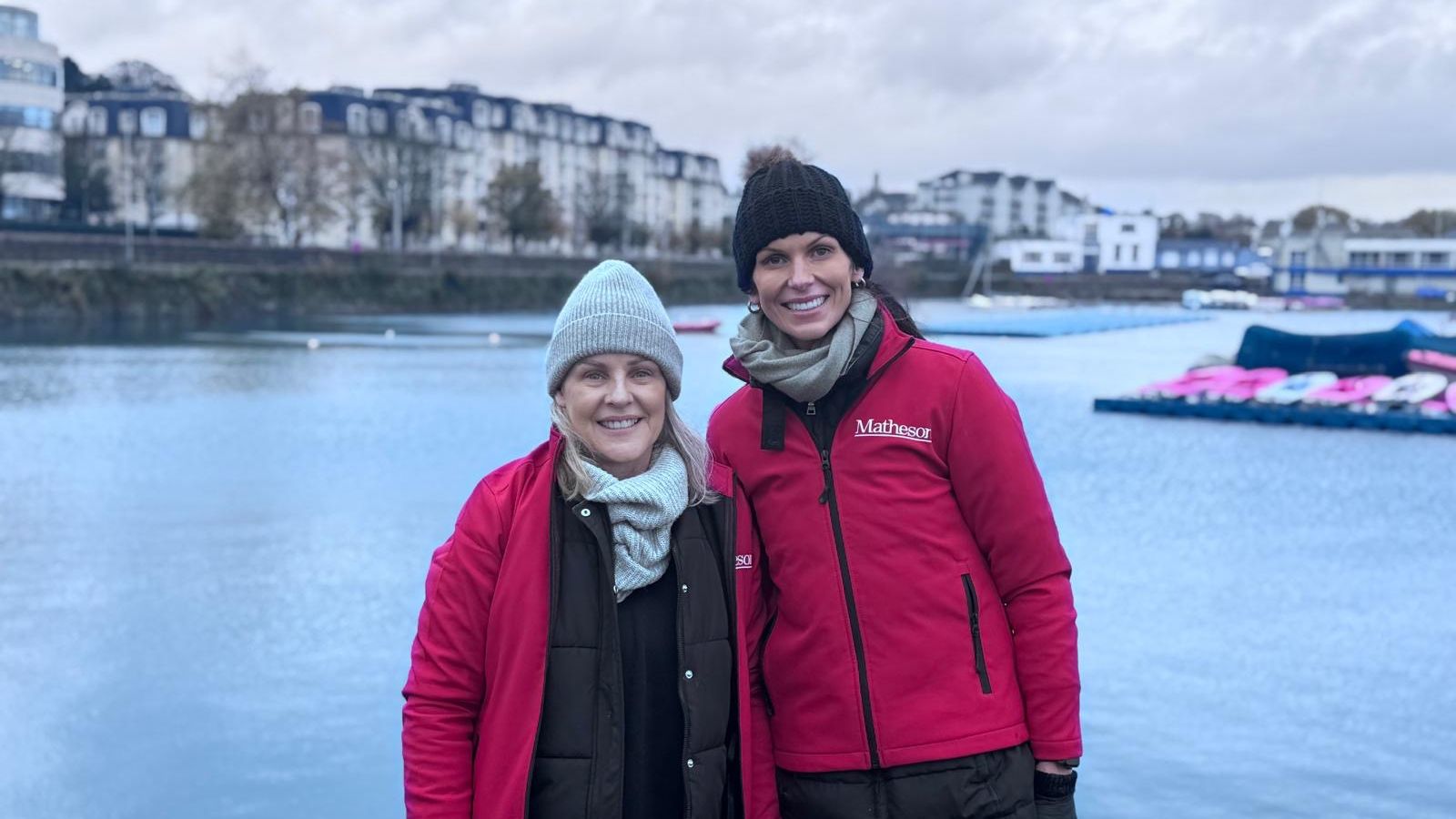 Two women wearing woolly hats and red jackets smiling in front of the marina water