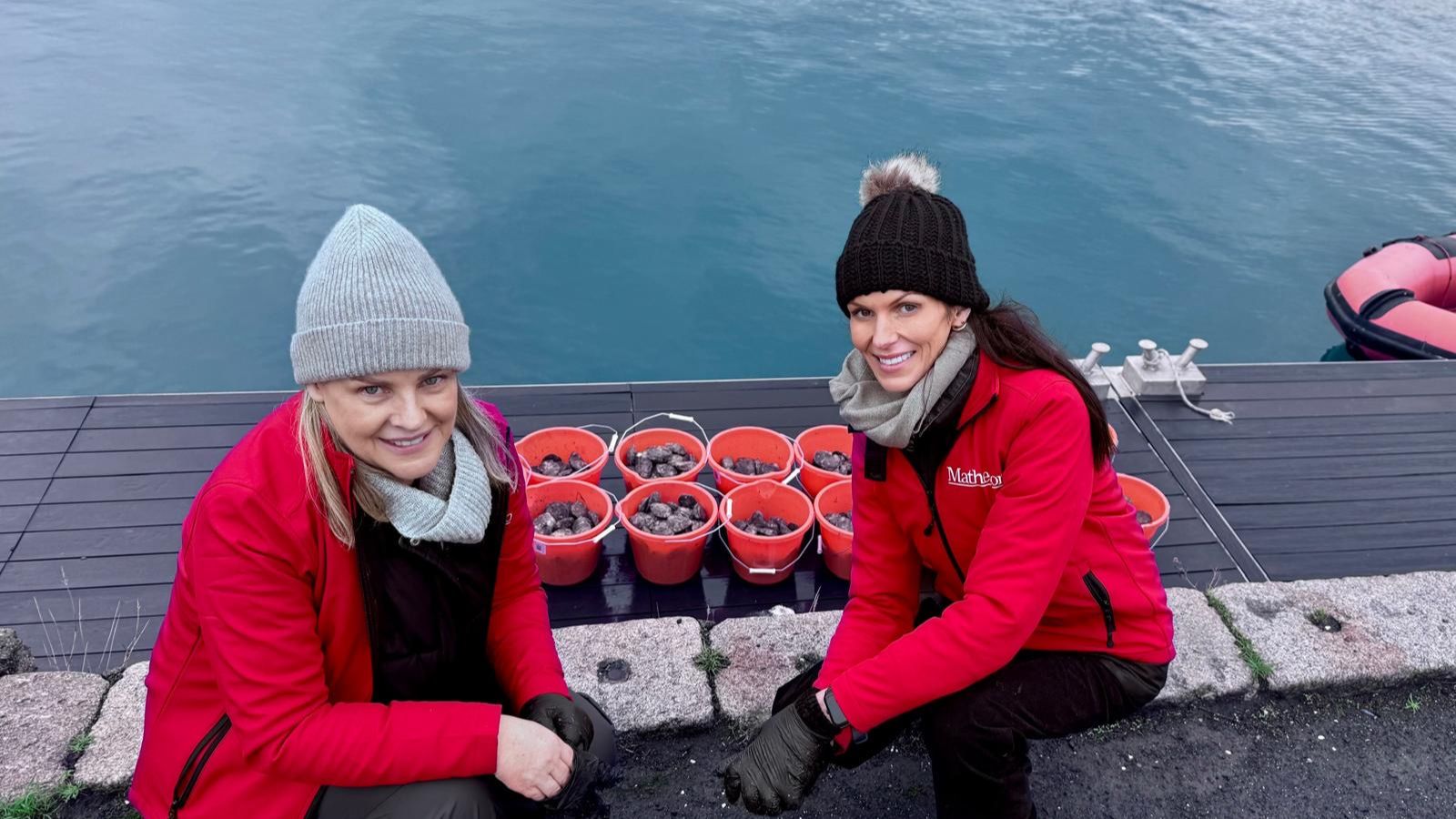 Two women wearing woolly hats and red jackets crouched down in front of orange buckets full of oysters by the marina water