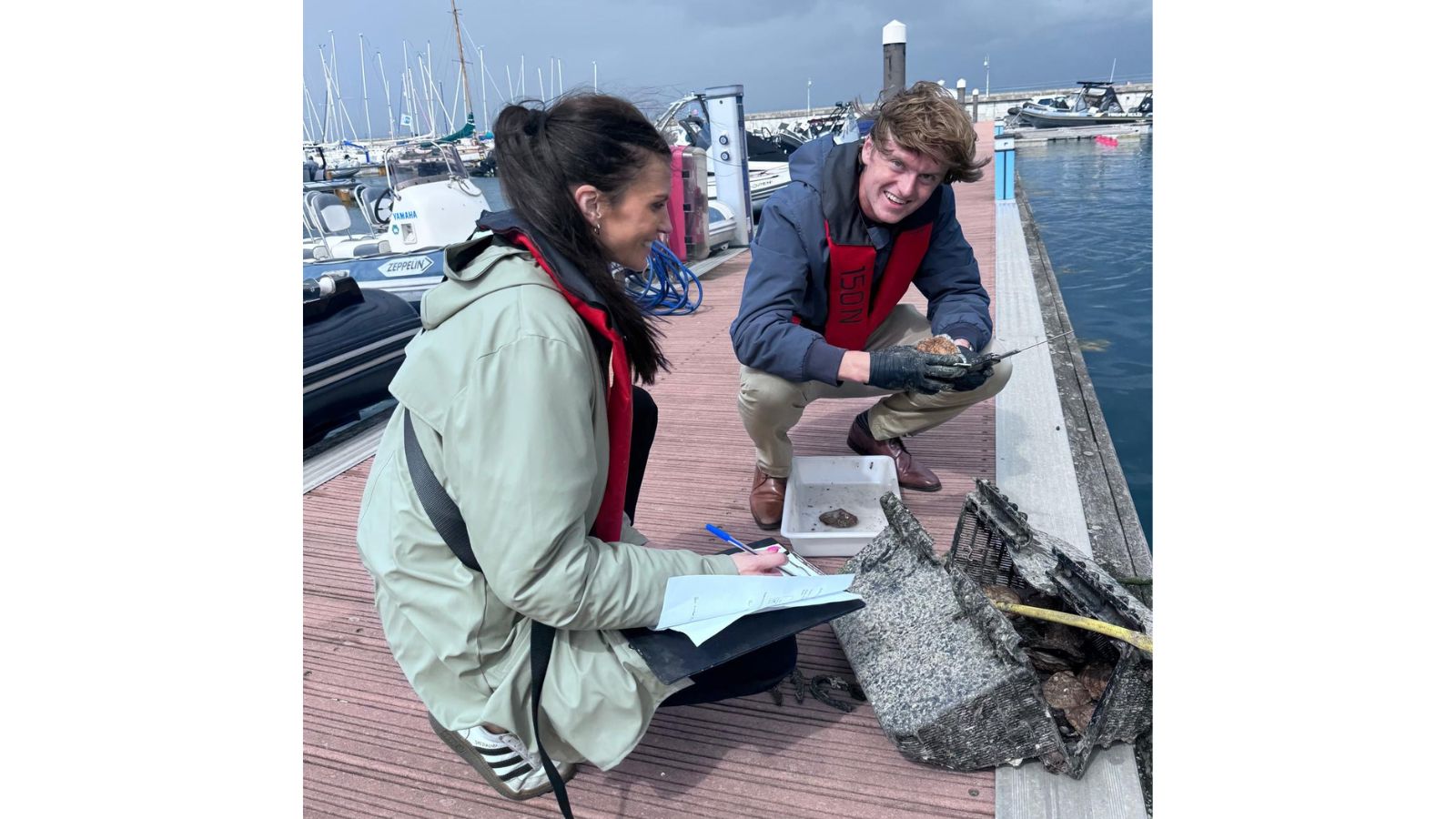 Two people smiling while writing down statistics and examining oyster baskets by the marina water